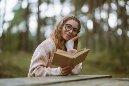 Happy woman is sitting at table with book in beautiful forest. Concept of relaxation, lifestyle.