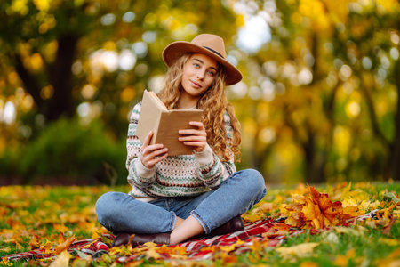 Beautiful curly woman in hat on mat with book in autumn park. Relaxation, solitude with nature.の写真素材