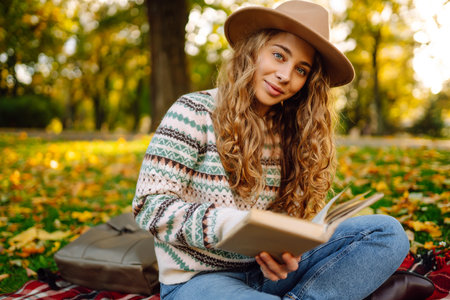 Beautiful curly woman in hat on mat with book in autumn park. Relaxation, solitude with nature.の写真素材