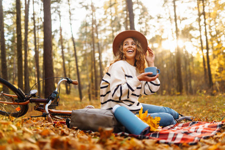 Hiker sitting on picnic blanket, drinking hot drink in autumn forest. Woman resting in nature.