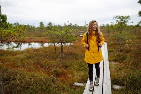 Smiling female traveler in yellow coat walks along wooden path among wild nature. Travel, vacation.の写真素材
