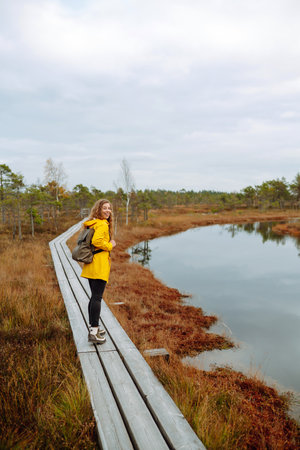 Smiling female traveler in yellow coat walks along wooden path among wild nature. Travel, vacation.の写真素材