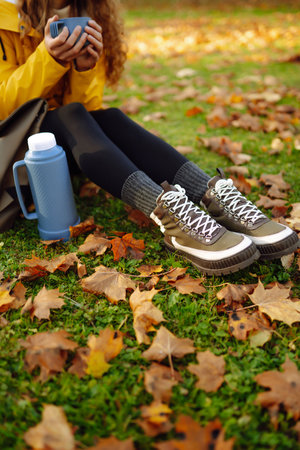 Woman traveler in yellow coat sits in clearing among yellow leaves and drinks hot drink from a flaskの写真素材