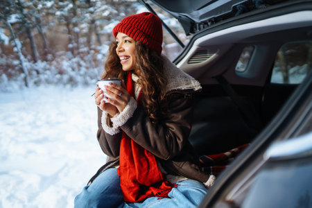 Happy woman holds a flask and drinks tea sitting in car trunk in winter forest. Lifestyle concept.の写真素材