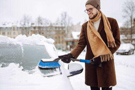 Stylish man in a coat and scarf clears snow from his car during winter snowfall. Seasonality conceptの写真素材