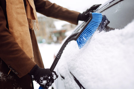 Stylish man in a coat and scarf clears snow from his car during winter snowfall. Seasonality conceptの写真素材