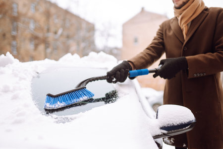 Stylish man in a coat and scarf clears snow from his car during winter snowfall. Seasonality concept