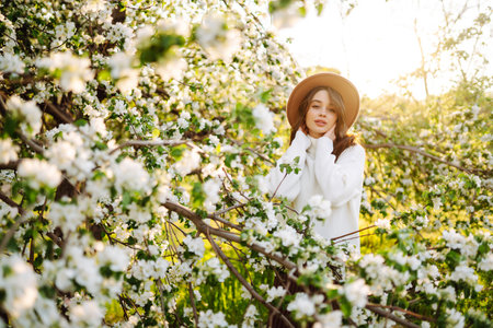 Portrait of Woman in hat posing near flowering tree. Smiling young woman enjoying the smell of...