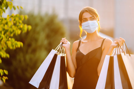 Fashionable woman wearing protective medical mask with shopping bags at sunset. Covid-2019.の写真素材