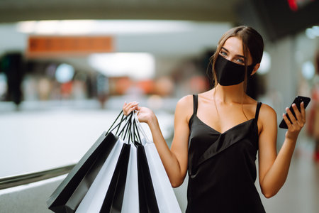 Young woman in protective black medical mask with shopping bags talking on a mobile phone in the mall.の写真素材