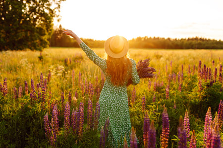 A young woman in a stylish green dress and hat enjoys the sunset in a field with lupins.の写真素材