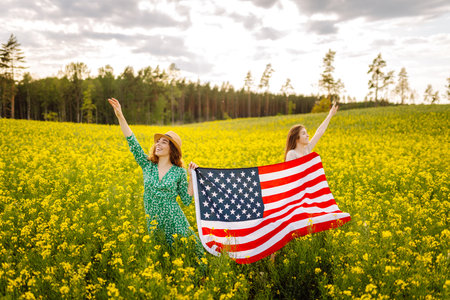 Two Young woman with american flag on blooming meadow. 4th of July. USA flag fluttering in the wind.の写真素材