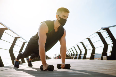 Handsome muscular man in protective mask, sports outfit doing functional exercises on seaside...