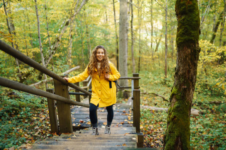 Woman in a yellow coat with backpack walks along wooden staircase, enjoying autumn scenery in forest