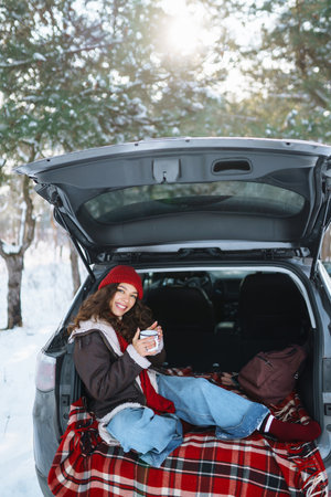 Young woman holds a thermos and drinks tea sitting in car trunk in sunny winter forest. Rest conceptの写真素材