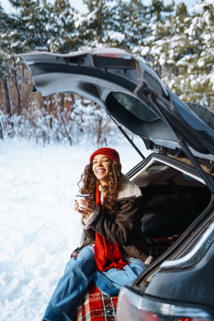 Young woman holds a thermos and drinks tea sitting in car trunk in sunny winter forest. Rest conceptの写真素材