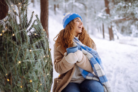 Young woman drinks a hot drink from a thermos while sitting in a snowy forest with a Christmas tree.