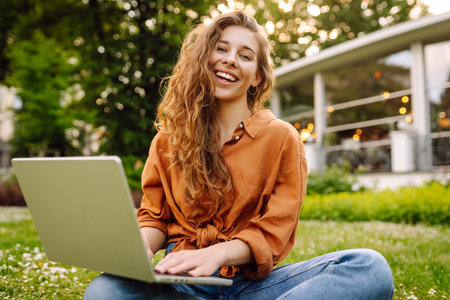 Beautiful female freelancer sits on green meadow with laptop. Online education. Freelancing concept.の写真素材