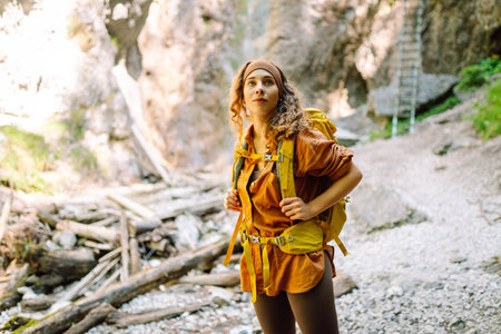 Smiling female traveler with yellow backpack enjoys waterfall near a mountain river. Nature concept.