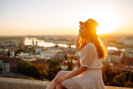Smiling female tourist in hat admires landscape of city at dawn. Euro-trip. Active lifestyle conceptの写真素材