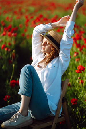 Young curly girl posing in a poppy field sitting on a chair. Summertime.の写真素材