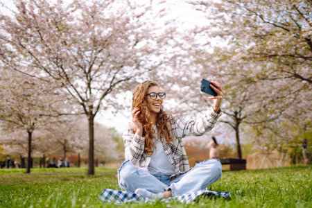 Young woman takes selfie against background of flowering trees in park. Concept of relaxation, fun.の写真素材