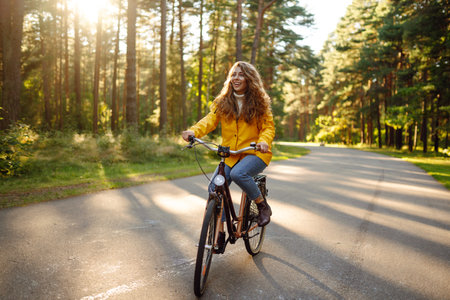 Young happy woman in yellow coat rides bicycle in sunny park. Woman enjoys autumn nature. Lifestyle.の写真素材