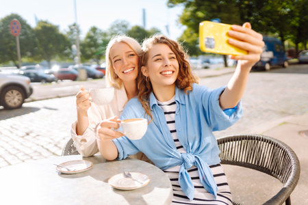 Young women taking selfie while sitting on terrace on sunny day. Girlfriends drink coffee with phoneの写真素材