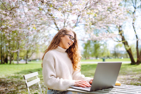 A young woman at a table in a blooming park works with a laptop. Freelancing, weekend concept.の写真素材