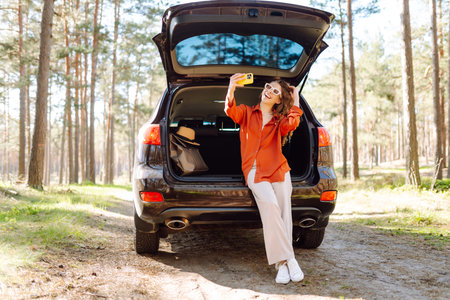 Happy woman sits on the trunk of her car and taking a selfie. Selfie while traveling by car.の写真素材