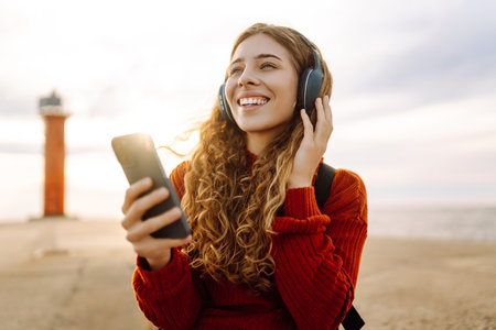 Happy woman listening nice music by the waterfront while wearing headphones by a lighthouse.の写真素材