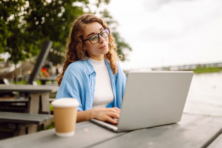 Portrait of a young woman in a blue shirt with a laptop at a table in an outdoor cafe near a lake.の写真素材