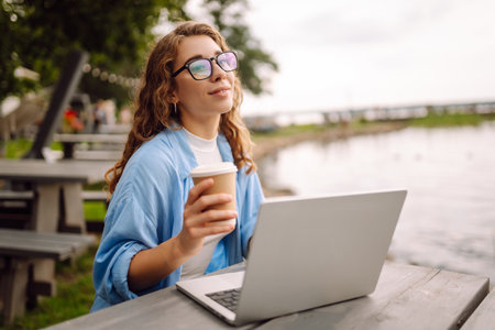 Portrait of a young woman in a blue shirt with a laptop at a table in an outdoor cafe near a lake.の写真素材
