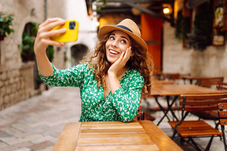 Young woman holding a smartphone in her hands and taking a selfie while sitting at the summer cafe.の写真素材