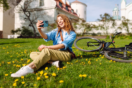 Young woman sits on green grass in a park and takes a selfie. Biking in nature. Relax and lifestyleの写真素材