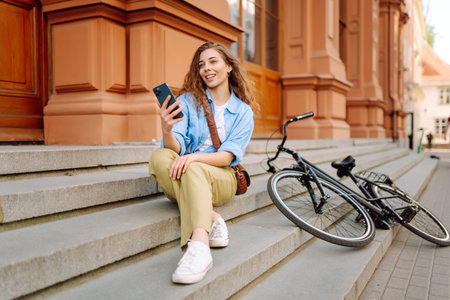 Young woman using mobile phone while sitting with bicycle at the city street outdoor.の写真素材