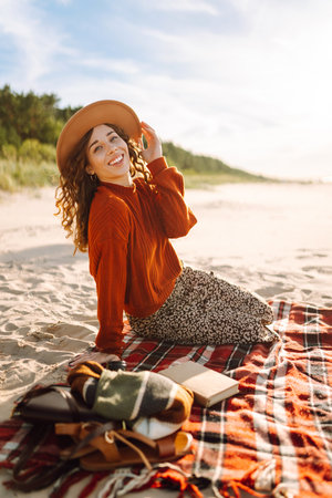 Young woman in cozy sweater, hat enjoys beautiful view of sea at beach picnic. Concept of relaxationの写真素材