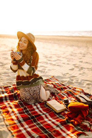 Woman sits on the beach drinking a hot drink from a thermos. Picnic, weekend, relax and lifestyleの写真素材
