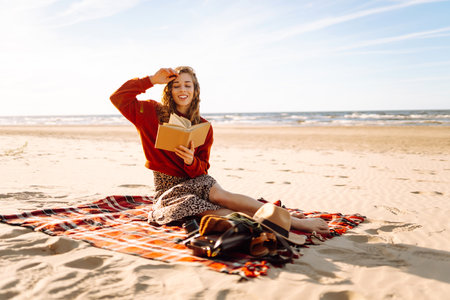 Beautiful woman sitting on blanket reading a book at beach picnic. Concept of relaxation, enjoyment.の写真素材