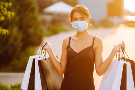 Fashionable woman wearing protective medical mask with shopping bags at sunset. Covid-19.の写真素材