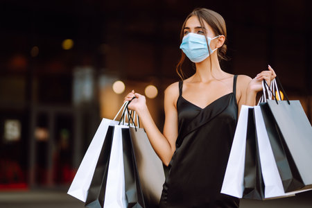 Fashionable woman in protective medical mask with shopping bags walking near mall. Covid-19.の写真素材