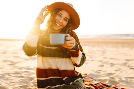 Cute woman sits on the beach drinks a hot drink from a thermos. Picnic, weekend, relax and lifestyleの写真素材