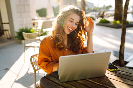 Young woman works at laptop on cafe terrace. Freelancer enjoying the weather outdoors. Vacation concept.の写真素材