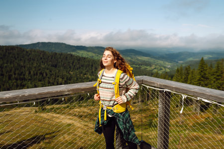 Happy woman with yellow backpack on wooden pedestrian bridge enjoying mountain scenery.の写真素材