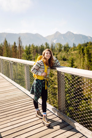Happy woman with yellow backpack on wooden pedestrian bridge enjoying mountain scenery.の写真素材