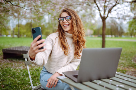 Cheerful woman on phone takes selfie while sitting at table with laptop in spring park. Lifestyle.の写真素材