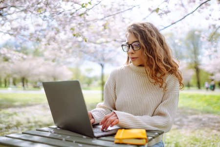 A young woman at a table in a blooming park works with a laptop. Freelancing, weekend concept.の写真素材