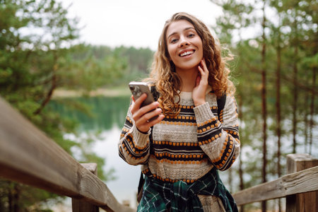 A young woman in a cozy sweater walking near a beautiful lake, smiling and using her phoneの写真素材