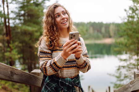 A young woman in a cozy sweater walking near a beautiful lake, smiling and using her phoneの写真素材