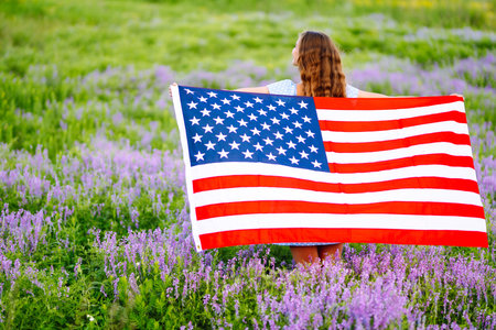 Young woman proudly hold waving american USA flag on blooming meadow. 4th of July. Independence Day.の写真素材